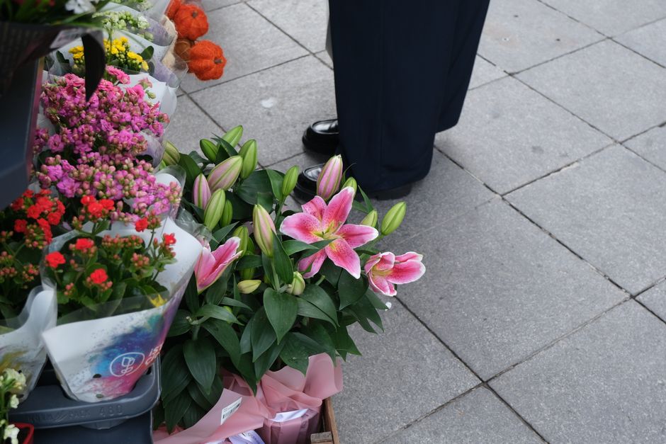 A floral arrangement featuring pink Asiatic lilies with large, vibrant petals, surrounded by dark green glossy leaves, positioned on a sidewalk at a flower stall. Nearby, there are other potted flower