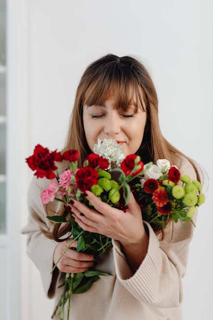 A woman with light brown hair and a beige sweater is holding a vibrant bouquet of fresh flowers close to her face, with her eyes gently closed and a slight smile. The bouquet includes bright red carna