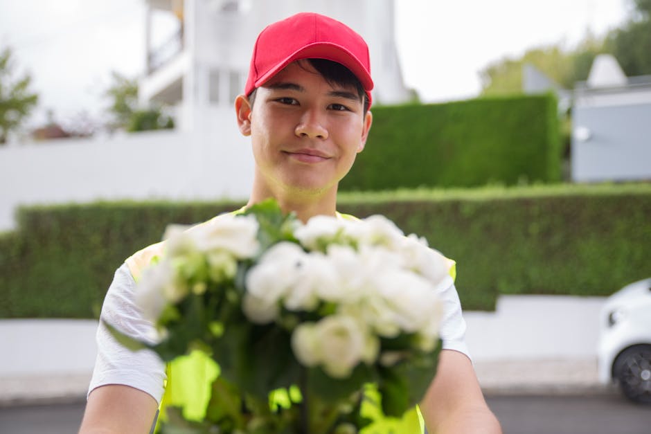 A delivery person wearing a bright yellow vest and a red cap is holding a bouquet of fresh white roses and green foliage, extending it forward as part of a flower delivery at an outdoor location. The 
