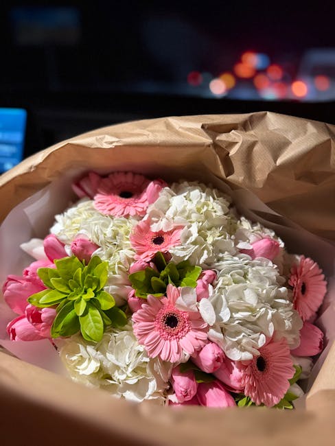 A bouquet of fresh flowers consisting of white hydrangeas, pink gerbera daisies, and green foliage, arranged in a natural style. The bouquet is wrapped in light brown craft paper with visible crumples
