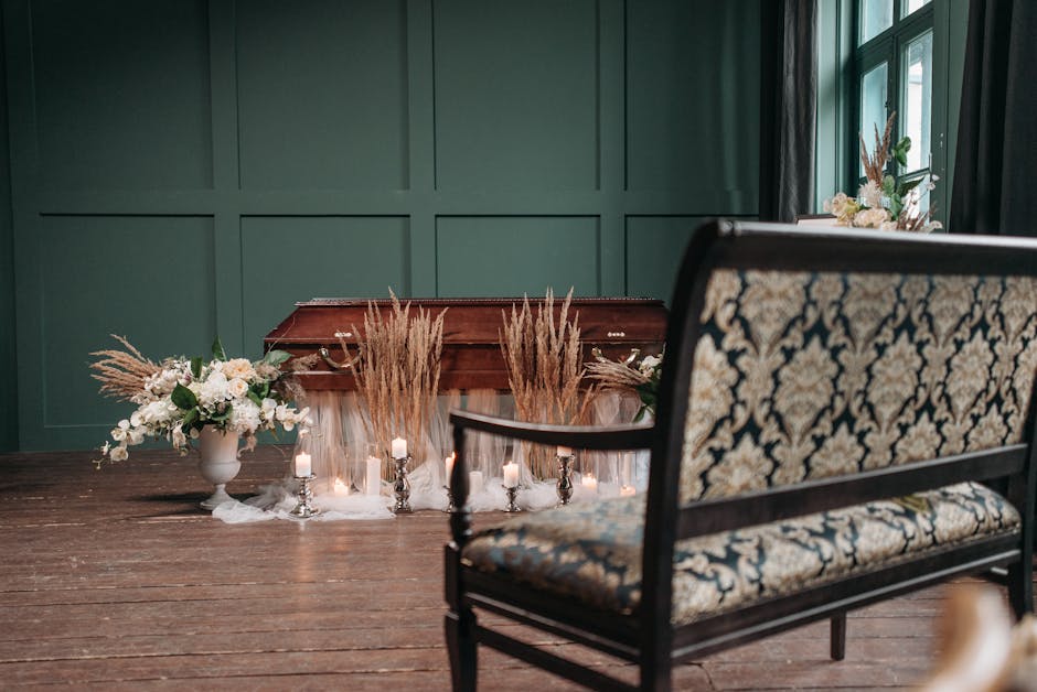 A floral arrangement featuring white and blush pink roses, white lilies, and soft greenery in a white ceramic vase, placed on a dark wooden table amidst tall, dried pampas grass and small lit candles,