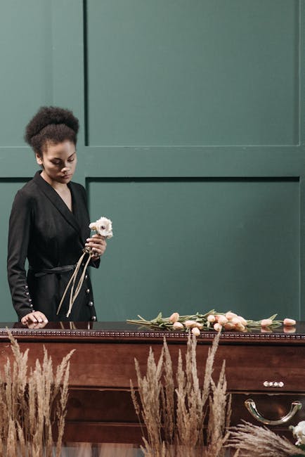 A woman with dark curly hair, dressed in a black blazer, stands beside a wooden coffin with a solemn expression, holding a small white flower bouquet. On top of the coffin, there is a floral arrangeme